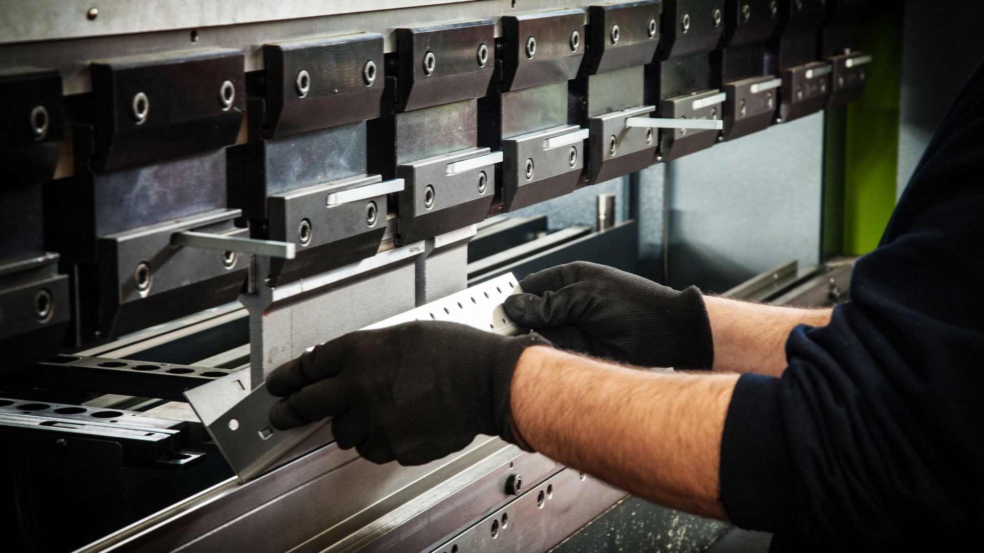 Gloved hands holding a punched metal part against the tooling of a press brake